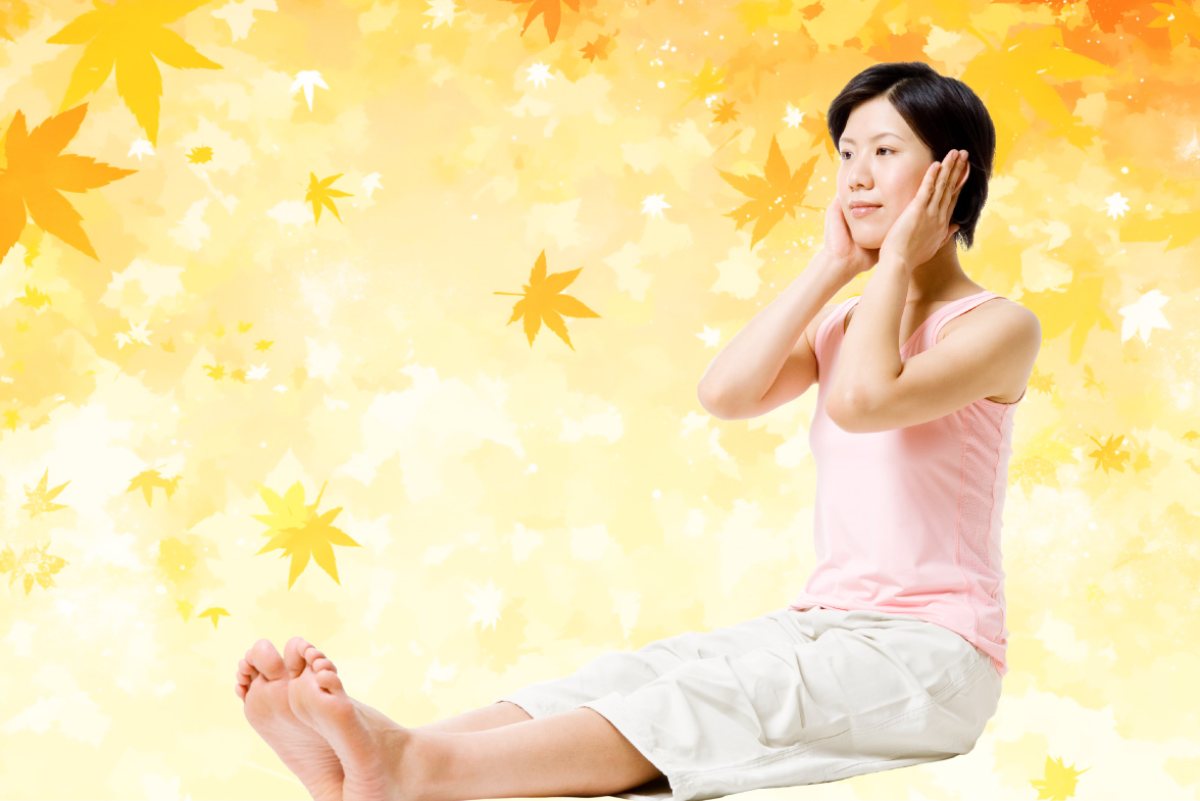 紅葉を背に、脚を伸ばして座り両手を耳にあてる女性